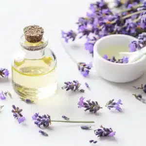 Een glazen flesje OsiMagnesium Good Night Melatonin Bodylotion (100 ml) met een kurk, omringd door paarse lavendelbloemen, staat naast een klein wit bakje met olie en een lepel.